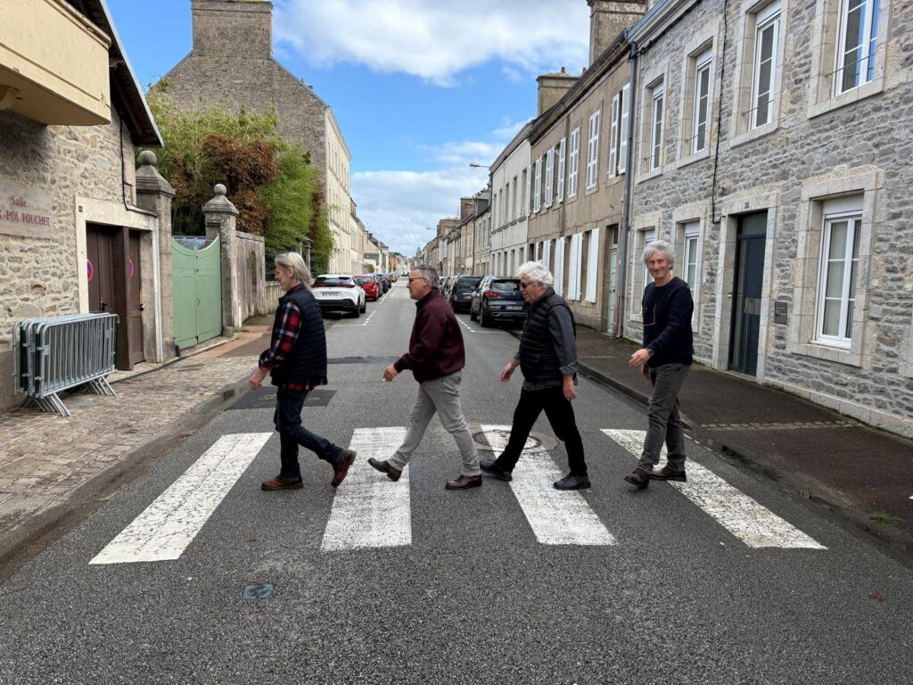 Quatre personnes traversent un passage piéton dans une rue calme d'une ville française, entourées de bâtiments en pierre et de voitures garées